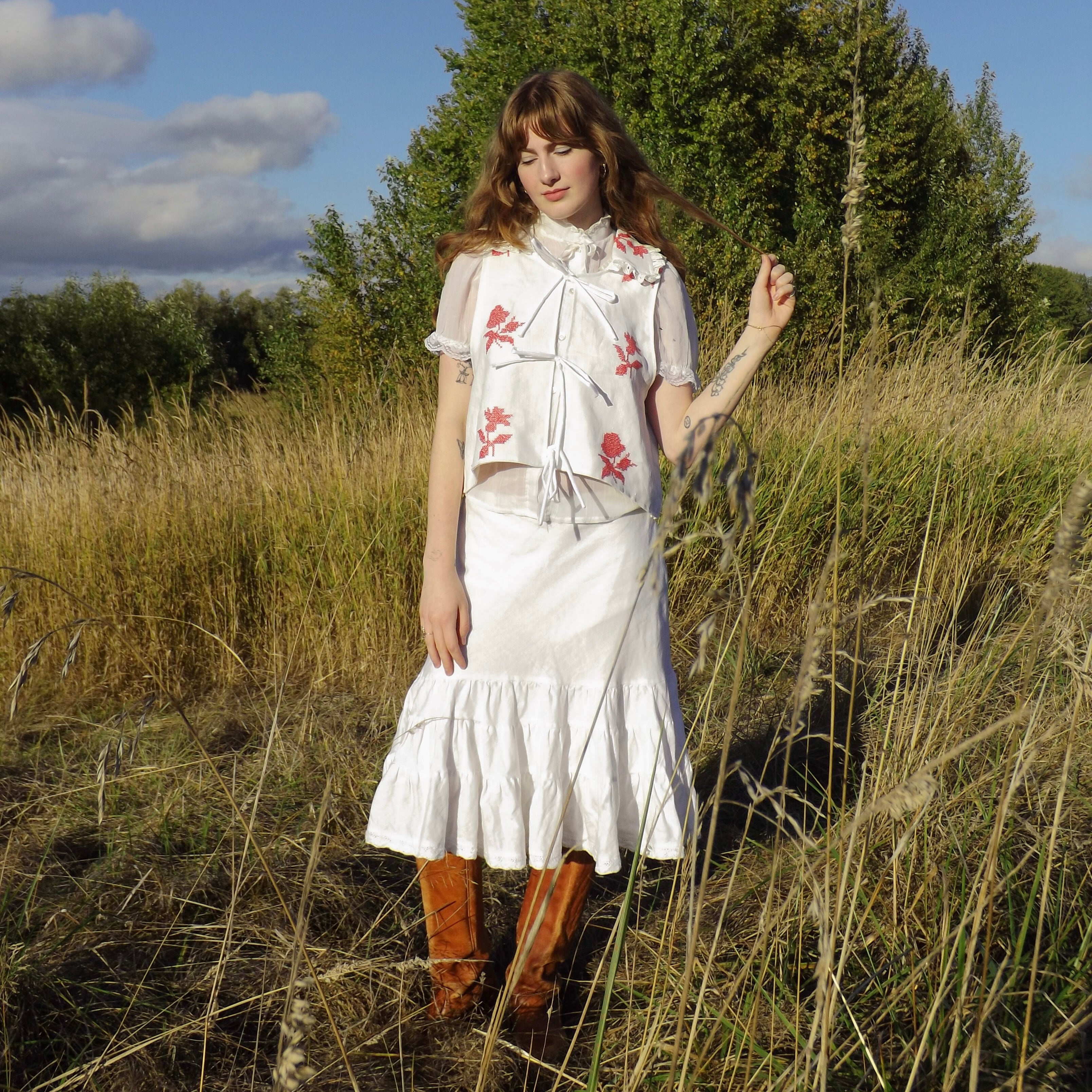 Woman in a white skirt, white embroidered tie-up blouse and tall boots standing in a field of tall grass. She's twirling a piece of her hair. 